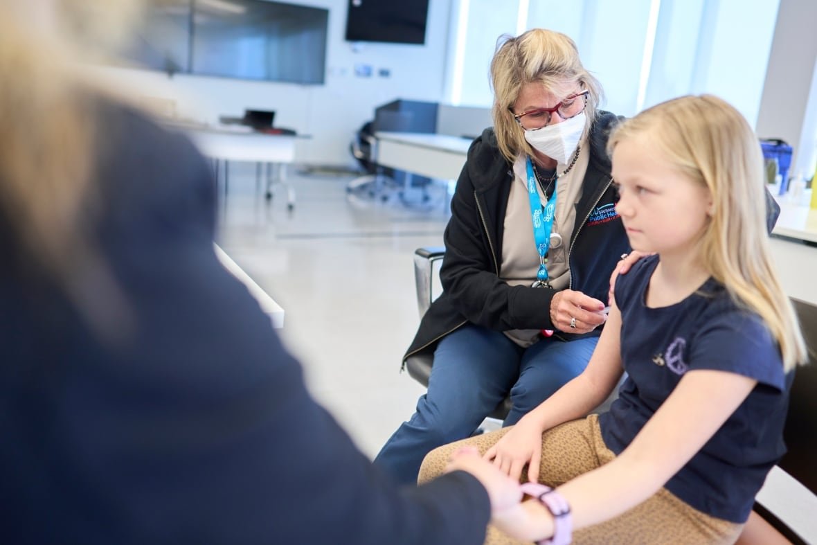 ont-measles-outbreak-20250306 A seated child holds the hand of a seated adult as a medical professional injects a needle into the child's arm.