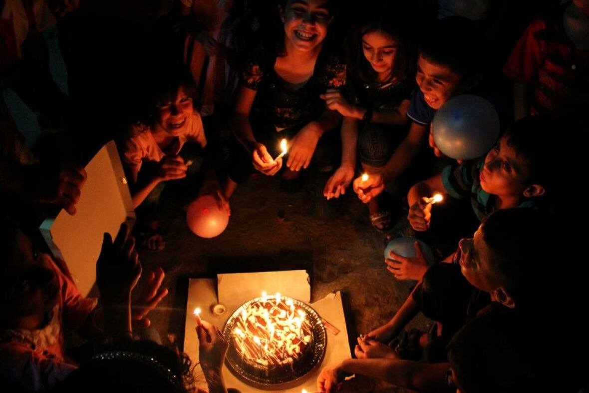 samar-abu-elouf-birthday-photo Children sit on the ground in a circle around a birthday cake, their smiling faces lit by the burning candles.
