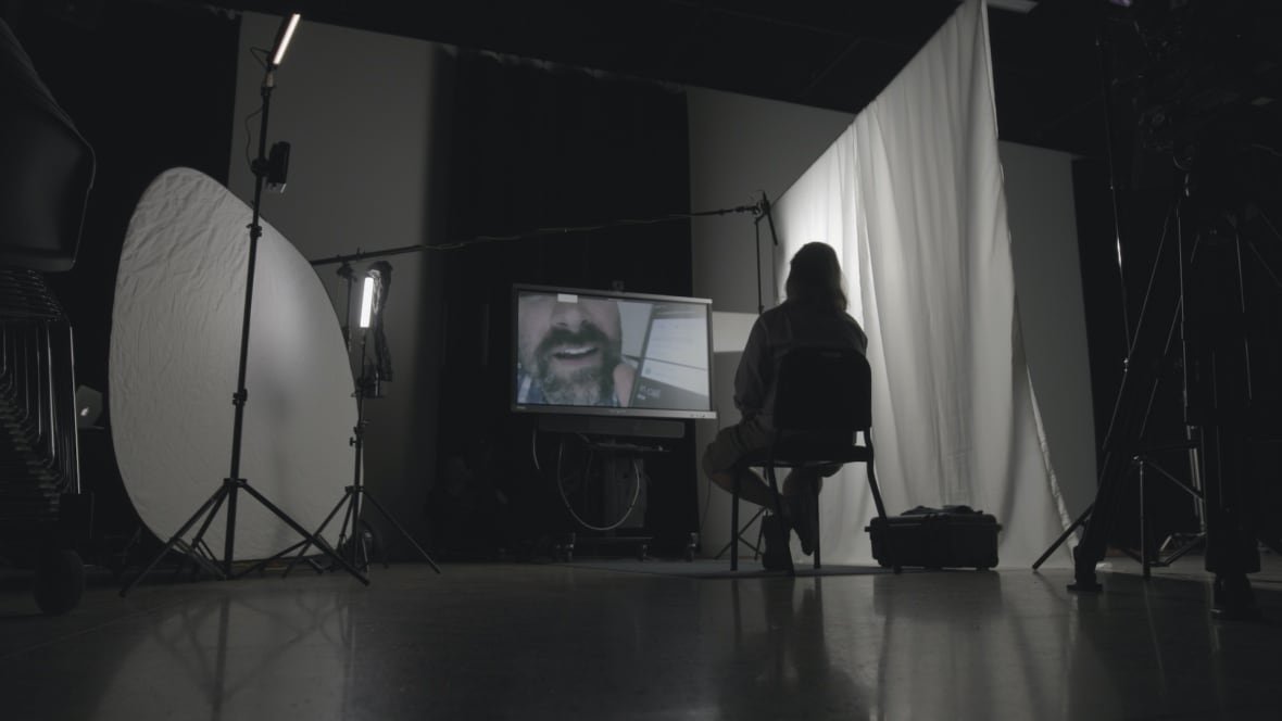 shamed A man sits in front of a screen with the projected face of a man mid-speech displayed on it.