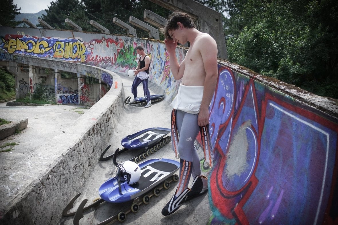 the-track-1 A man wearing a skin-tight aerodynamic suit stands on a concrete luge track. At his feet is a luge sled on wheels.