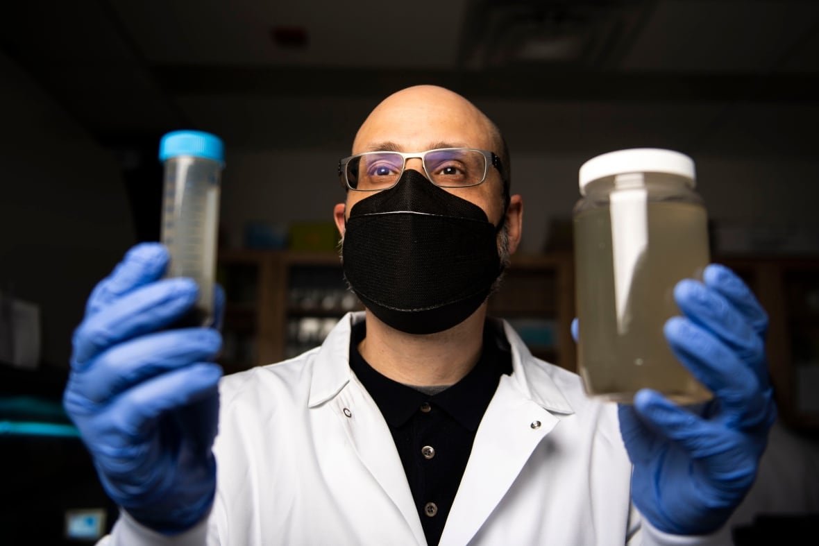 wastewater-testing A man in a facemask holds up a jar and a test tube with murky liquids inside them.