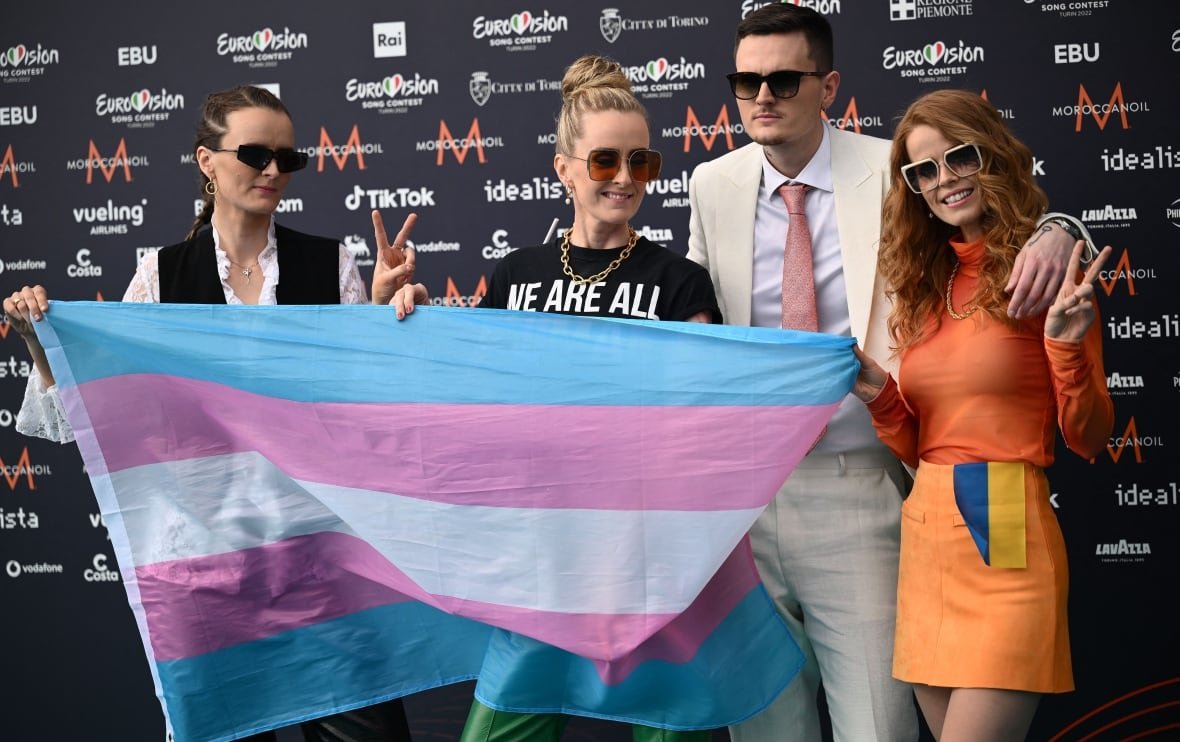 1240537708 Four people stand, three of them holding up a large transgender flag. The woman on the far right is in an orange outfit and has the Ukraine flag tucked into her skirt.