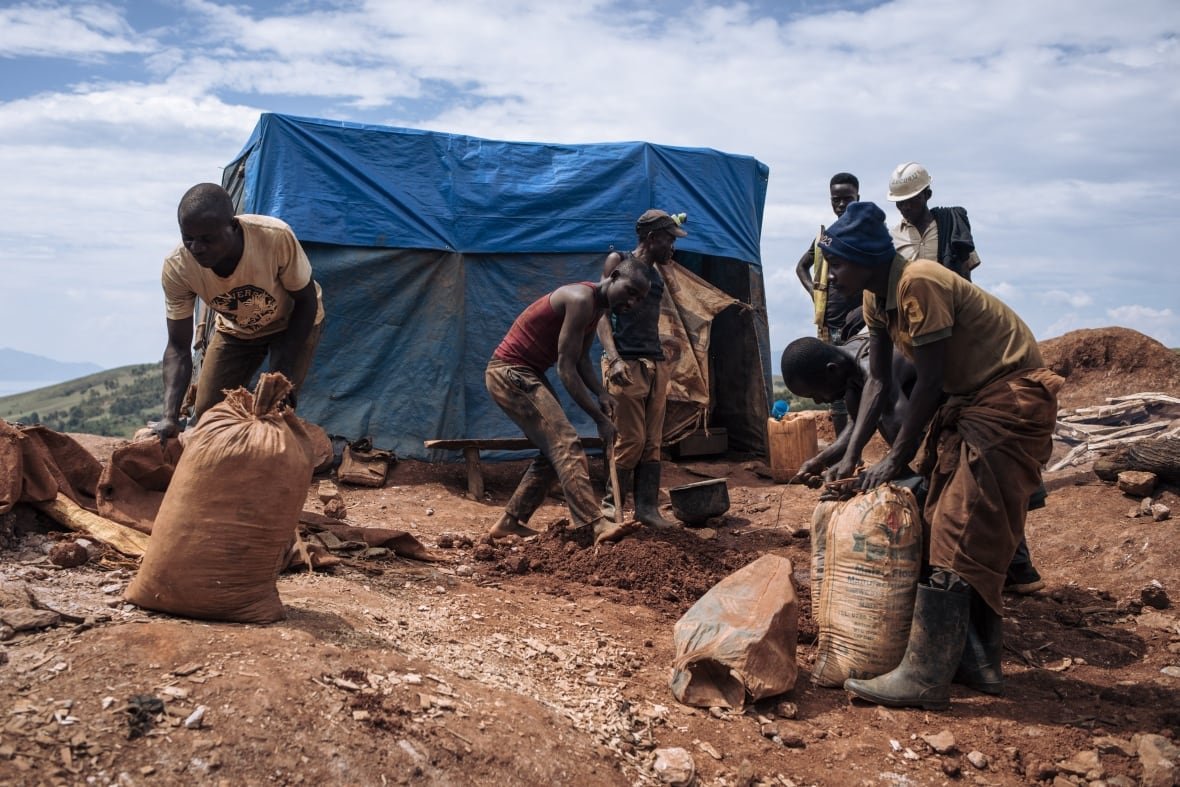 1256045330 About a half dozen dark complected men toil on dirt ground, filling large bags and working with shovels.