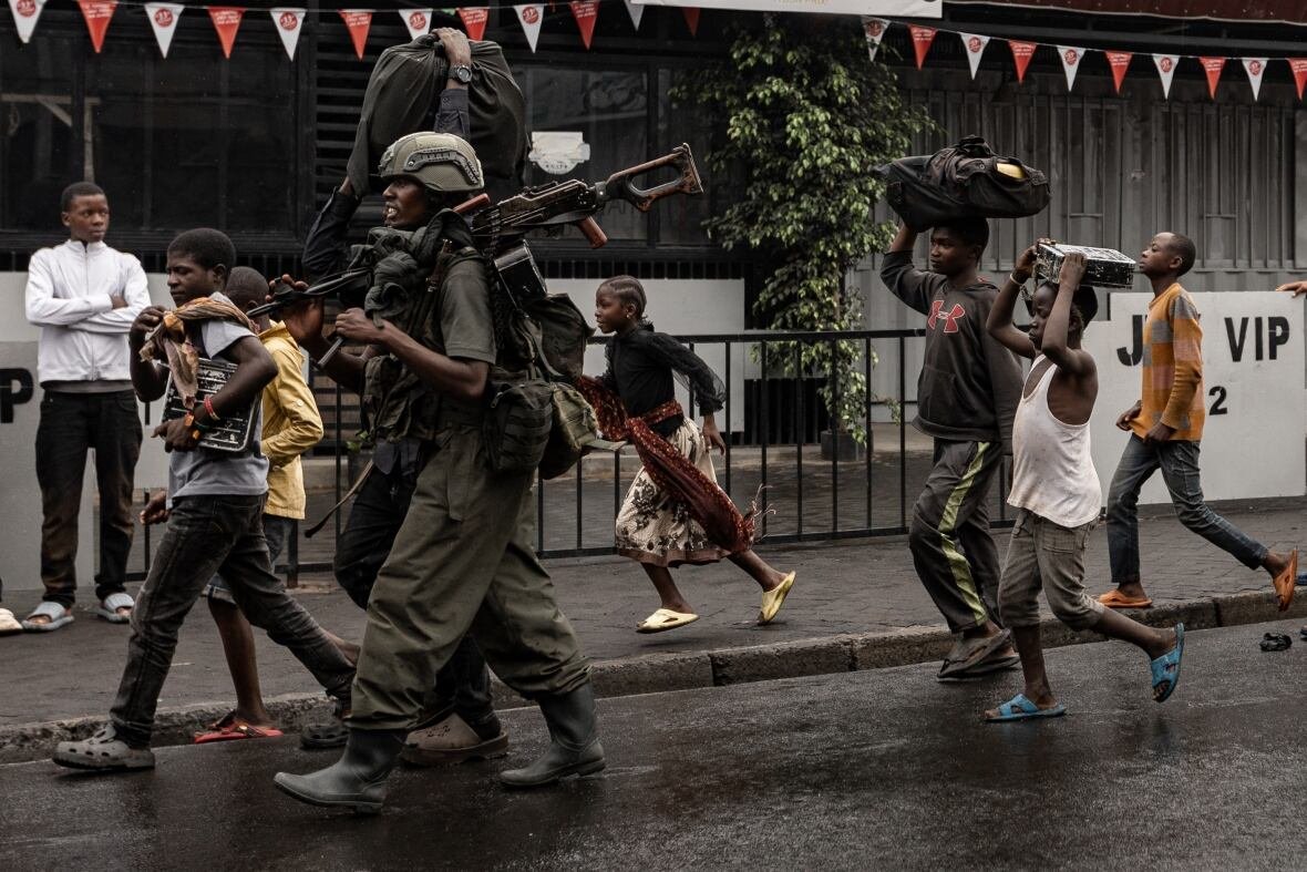 2195682067-1 A helmeted man with a large gun strapped on his fatigues is shown walking down the street surrounded by other dark complected people, civilians which include children.