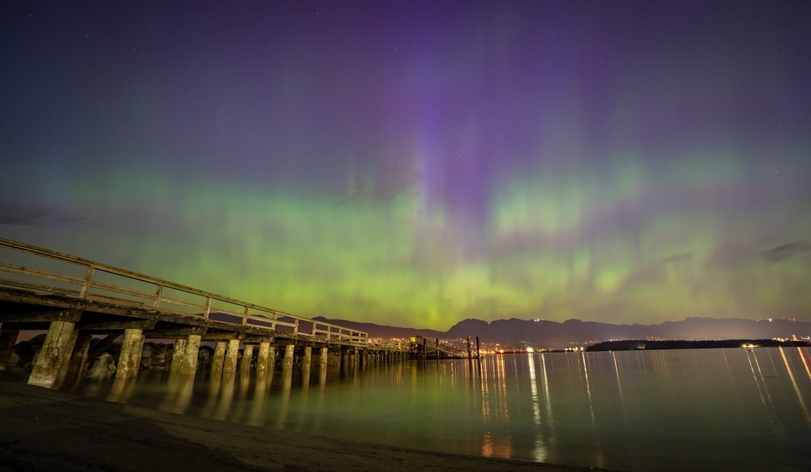 aurora-borealis-vancouver-20240511 Northern lights are seen above a bridge and water, with mountains visible in the distance.