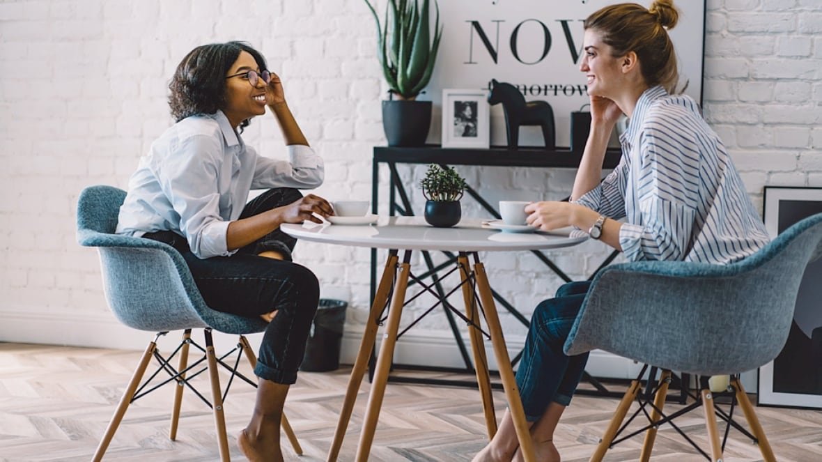 awkward-friends Two smiling women sit opposite each other at a table, drinking coffee, in a living room.