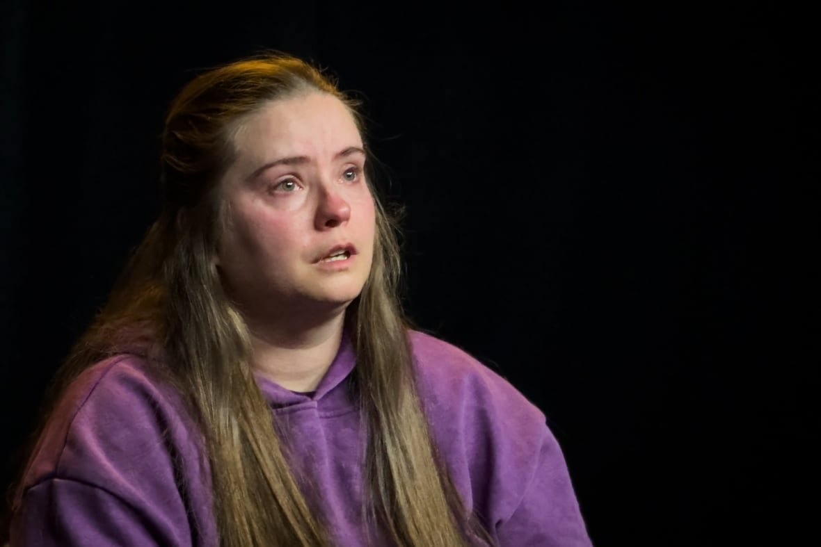 carley-gibbs A woman with long hair in a purple top speaks while looking sad.