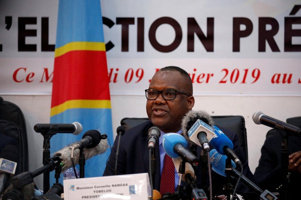 congo-security-nangaa-pix A cleanshaven, dark complected man with glasses and wearing a suit and tie sits at a table and speaks with a banner and flag on the wall behind him.