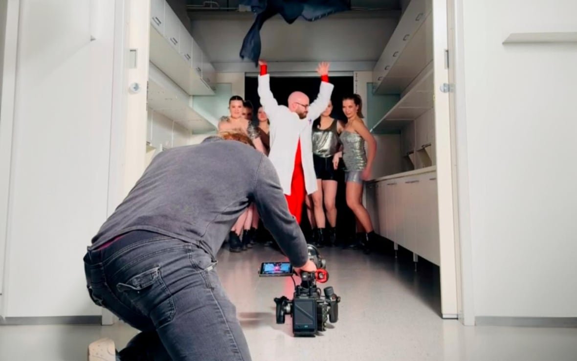 dance-your-phd A man kneels on the floor, facing away from the camera, pointing a video camera at a blad man in a bright red suit and labcoat standing in front of a group of women in sparkling disco dresses.