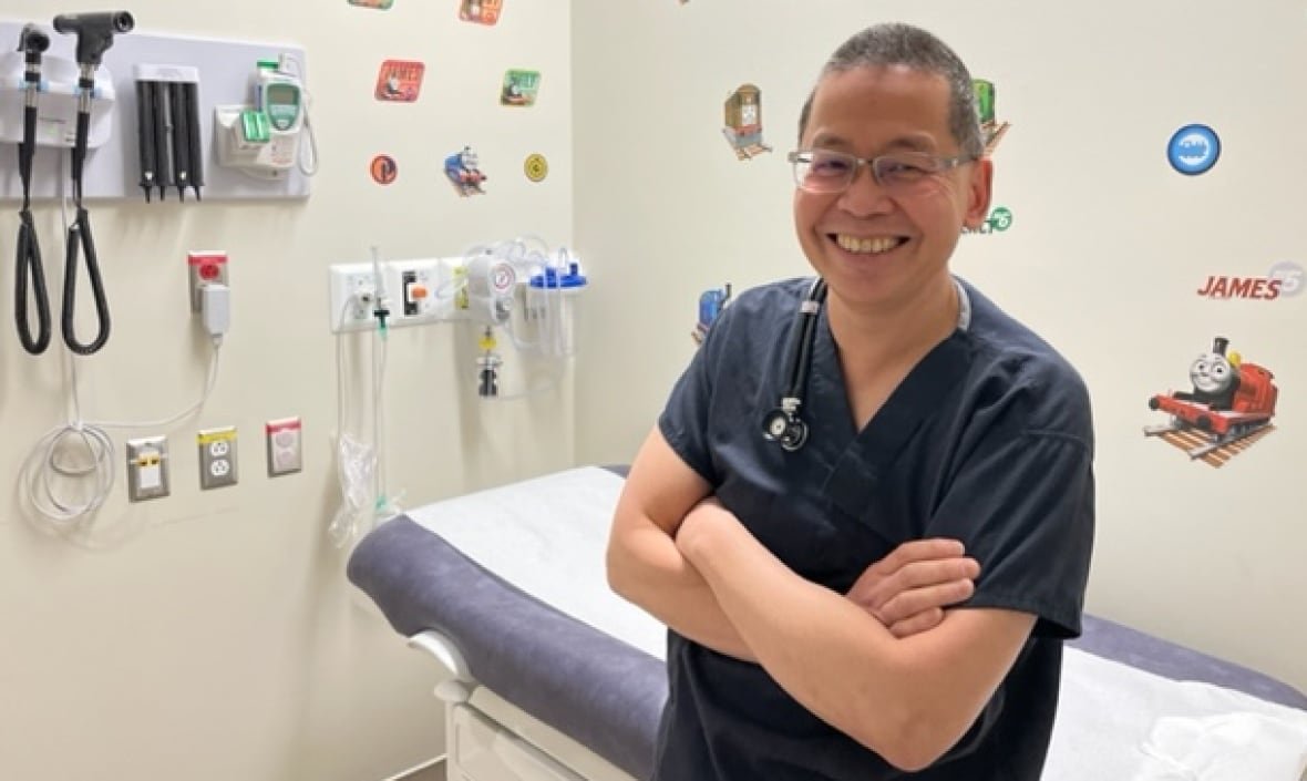 dr-sam-wong-alberta-medical-association-pediatrician A smiling pediatric doctor in blue scrubs is seen in an unoccupied patient's room adorned with colourful stickers on the walls.