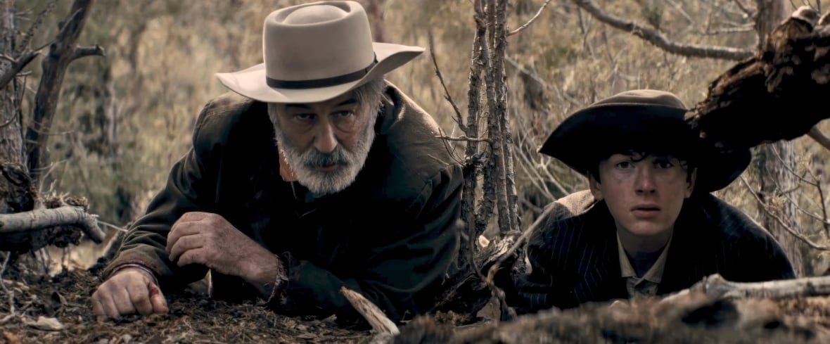 film-review-rust-1 An older man in a cowboy hat and a teenage boy lie on their stomachs on the ground in a forest, looking towards the camera and beyond it.