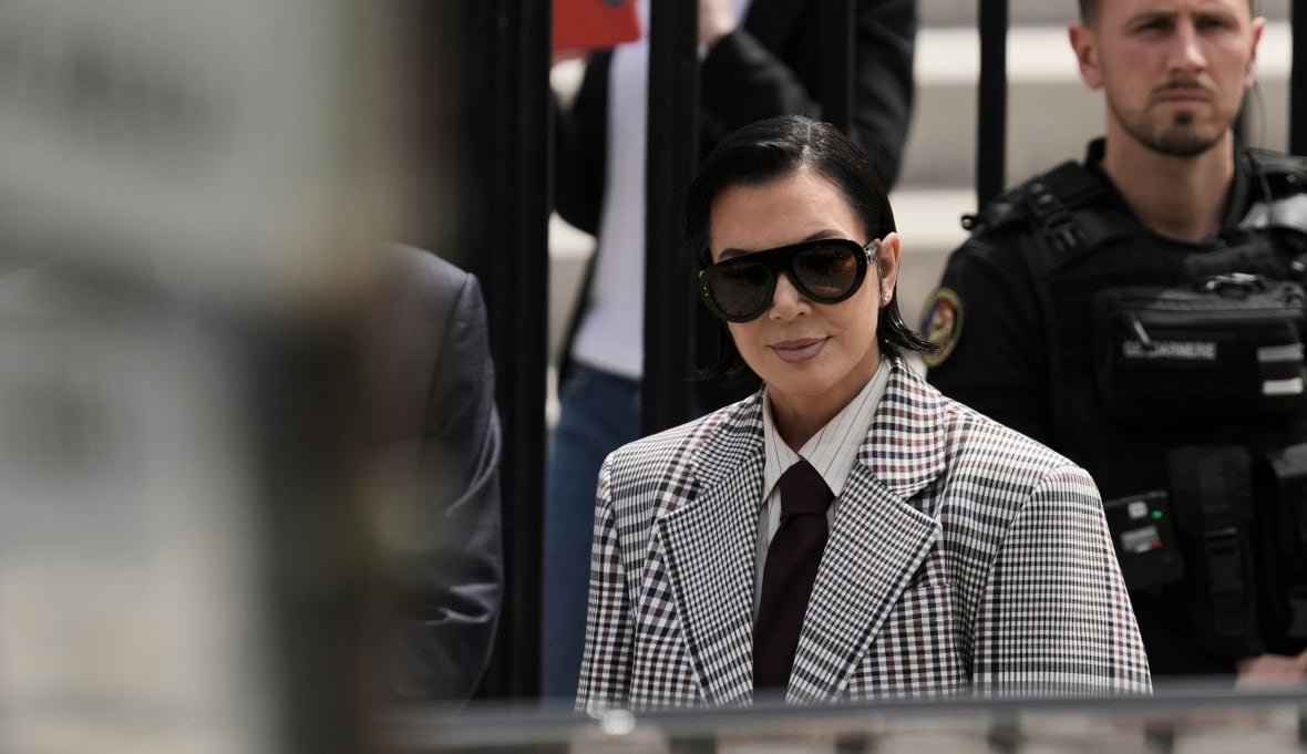 france-kardashian-robbery-trial A woman in a white and black plaid suit jacket is shown from the waist up. An officer is visible standing behind her, and the photo appears to be taken from a distance, with a blurry fence or barrier close to the camera blocking the left half of the image.