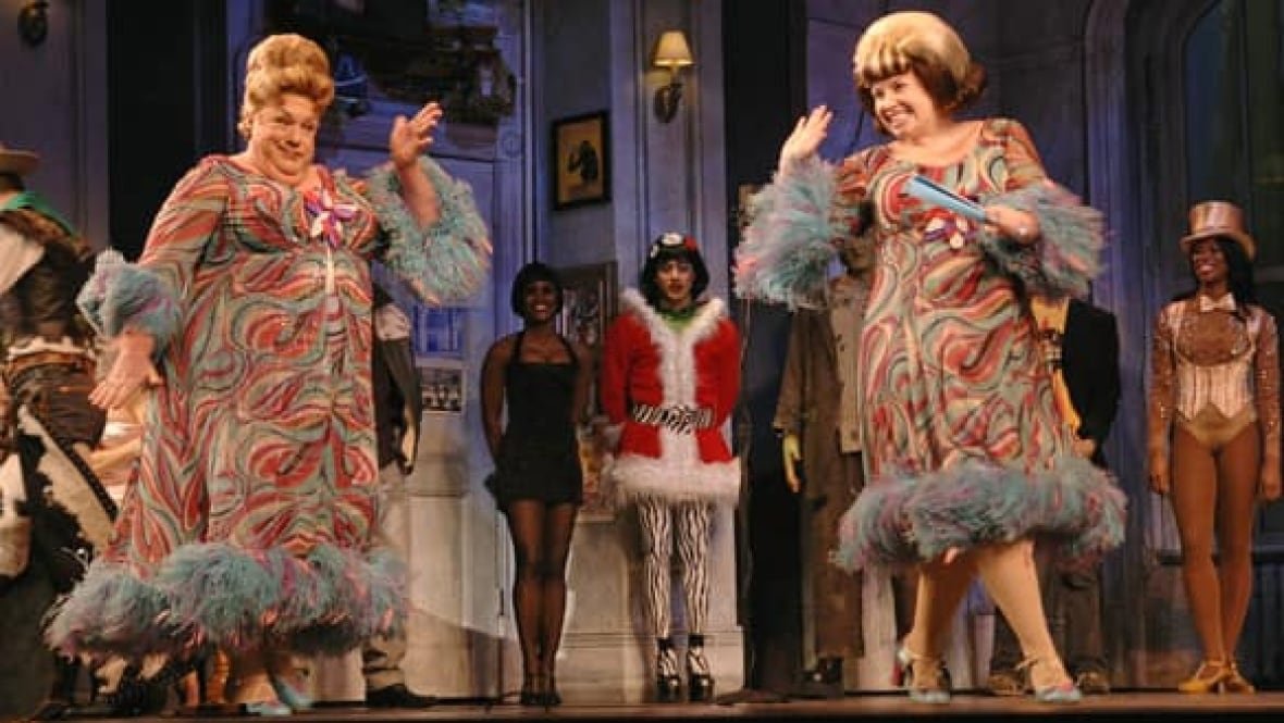 hairspray-wendt A man wearing a very loud print dress and large wig performs on stage next to a woman in a similar outfit.