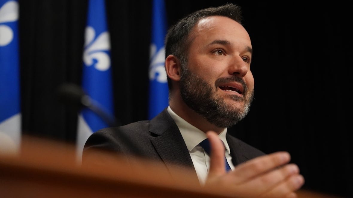 mathieu-lacombe Man speaking at podium with three Quebec flags behind him.