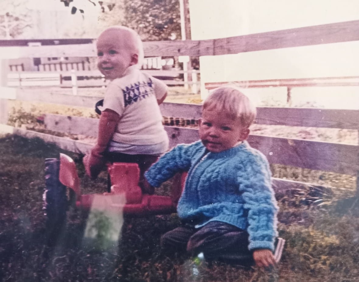 richard-anderson-first-person-bruce A faded photo shows two little boys beside a fence in a yard. One is sitting on a red toy tractor and looking to the left, the other is sitting on the ground looking directly at the photographer.
