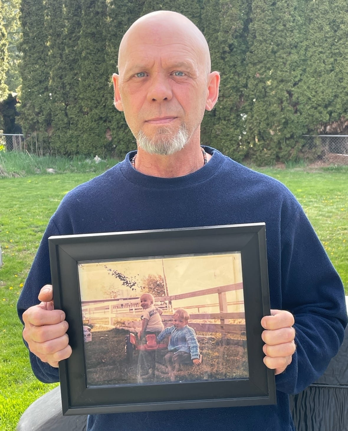 richard-anderson-first-person-with-photo-of-brother-1 An unsmiling man holds a framed photo showing two young boys playing in a yard.