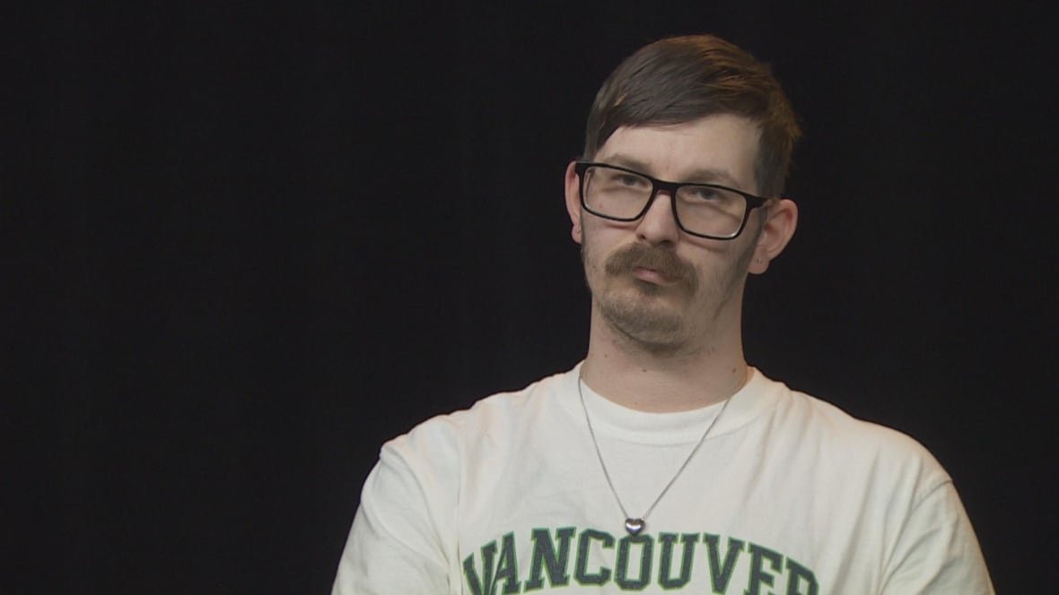 tyler-gibbs A young mustachioed man with short hair in a white top that says Vancouver in green looks at the camera with a serious expression.