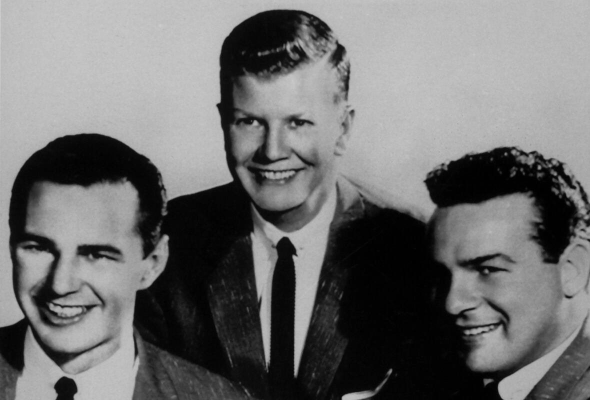 1153739812 Three men in suits pose with the man in the centre standing higher behind the other two men, who are seated.