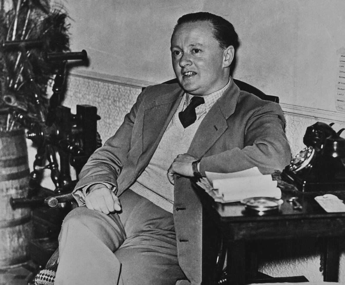 2150814198 A black and white photo shows a man in a suit and sweater vest sitting in a chair with one elbow resting on a small desk next to him.