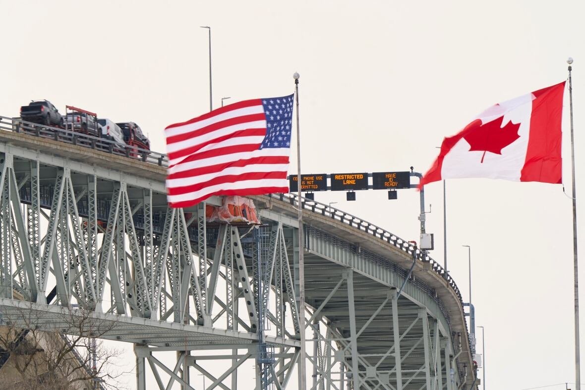 2195990033 The Canada and U.S. flags fly in the foreground as vehicles travel on a bridge in the background.