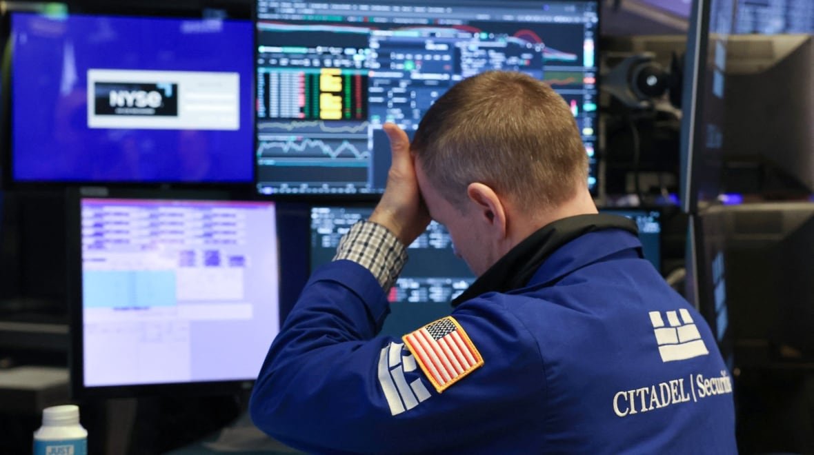2208435568 A man, sitting in front of several computer monitors, puts his hand to his forehead, as if in exasperation.