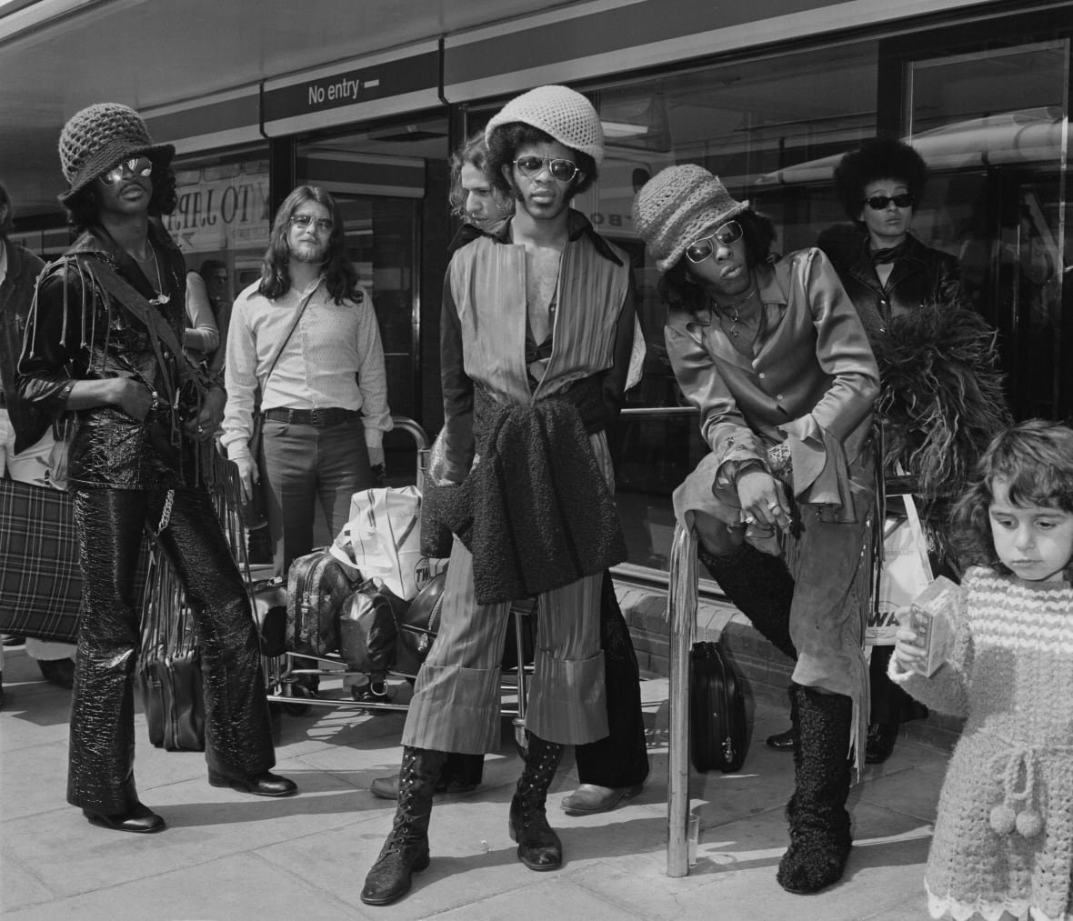 In this black and white photo, five people pose. The central person is looking at the camera and is slightly in front of the group, with someone else sitting on a table and others standing around. A small child passes by the corner of the image, seemingly unaware of the camera.