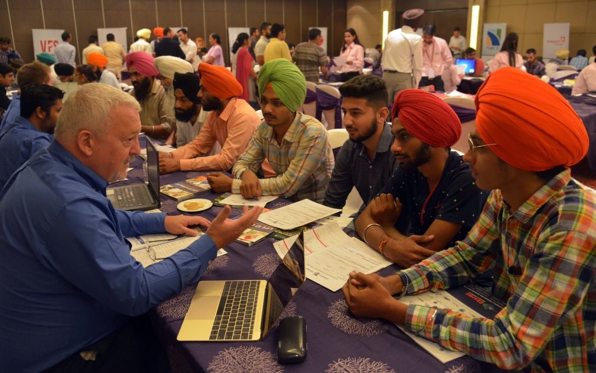 An adult sitting on one side of a table talks to several younger adults sitting on the other side of the table.
