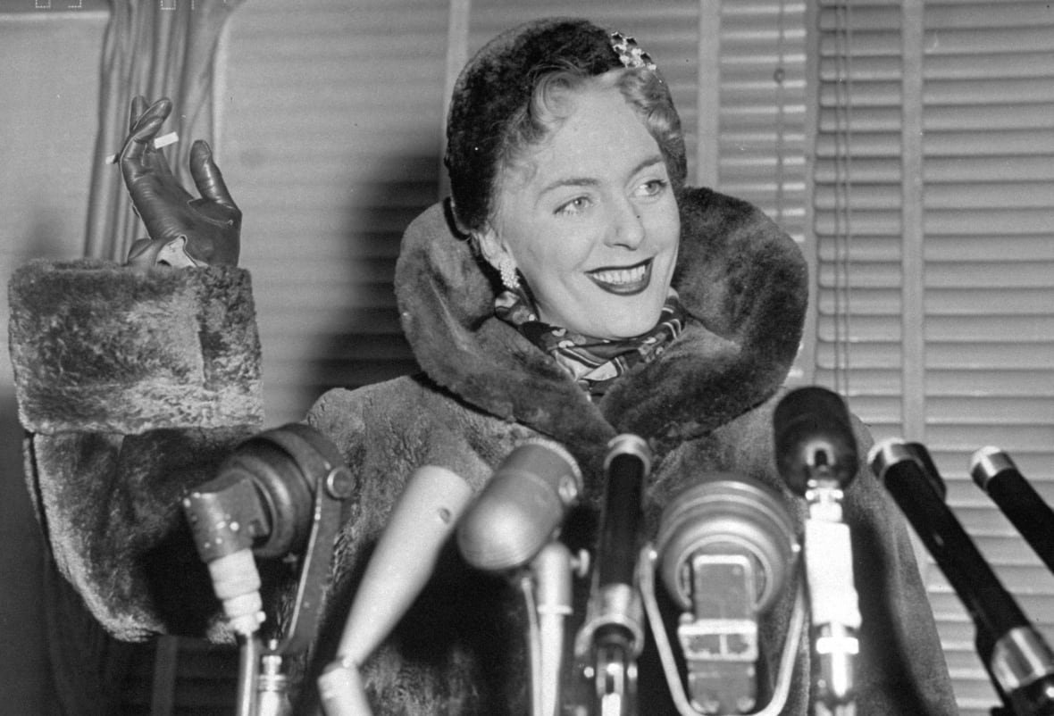 97258116 In this black and white picture, a smiling woman stands in a fur coat in front of a row of microphones. She has a cigarette in her raised, gloved hand.