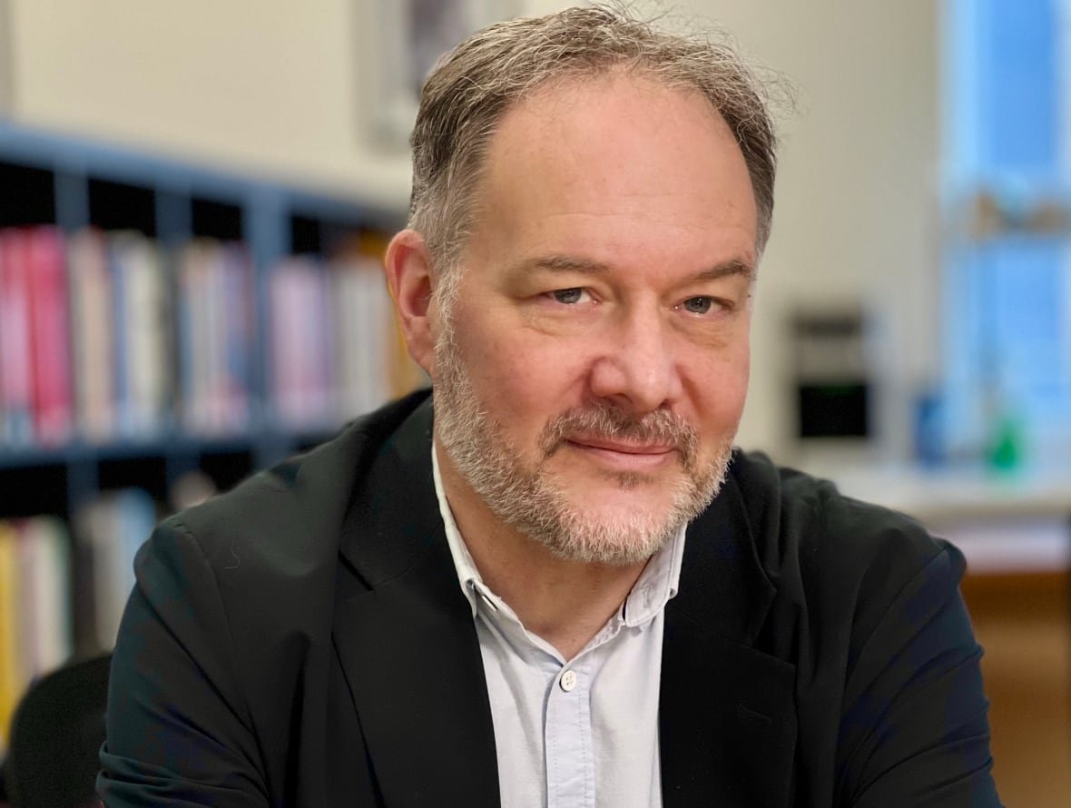 A close-up portrait of a man in a blazer and button-down shirt sitting indoors, with bookshelves and a window seen behind him.