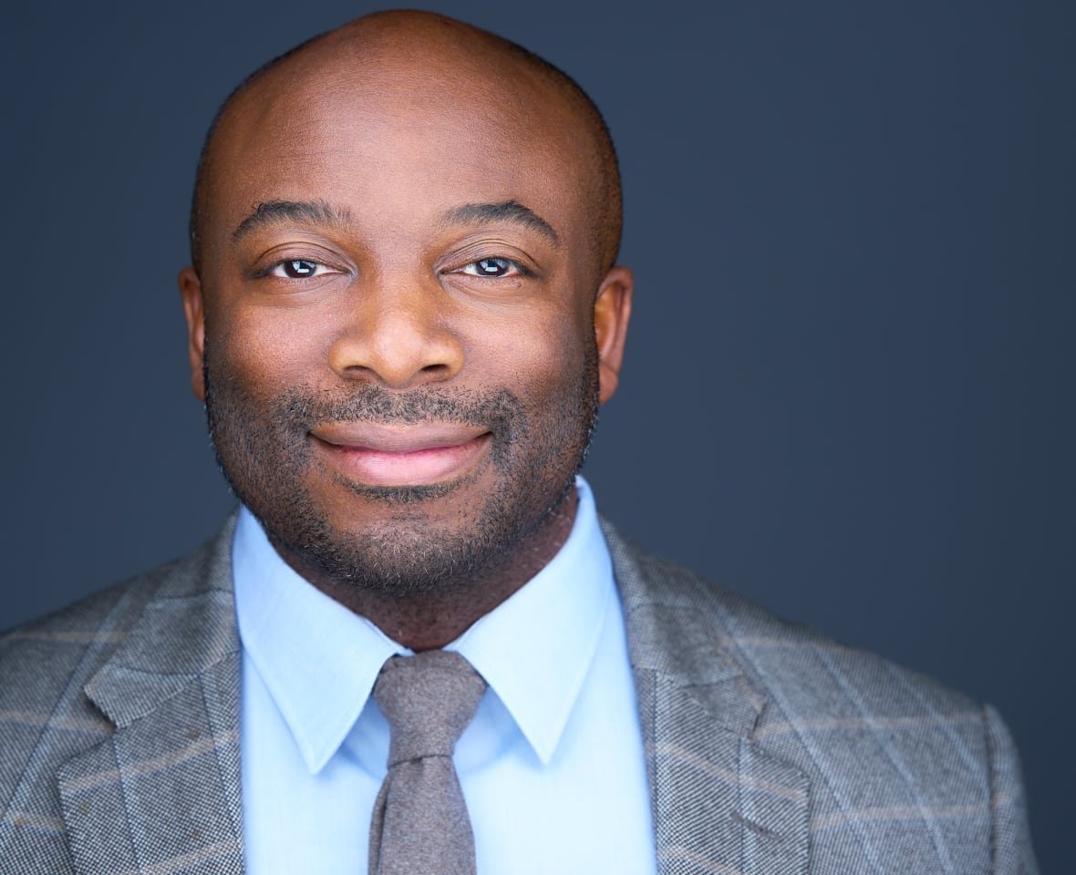 A man with a shaved head and slight beard smiles while wearing a grey, plaid suit and tie with a blue shirt.