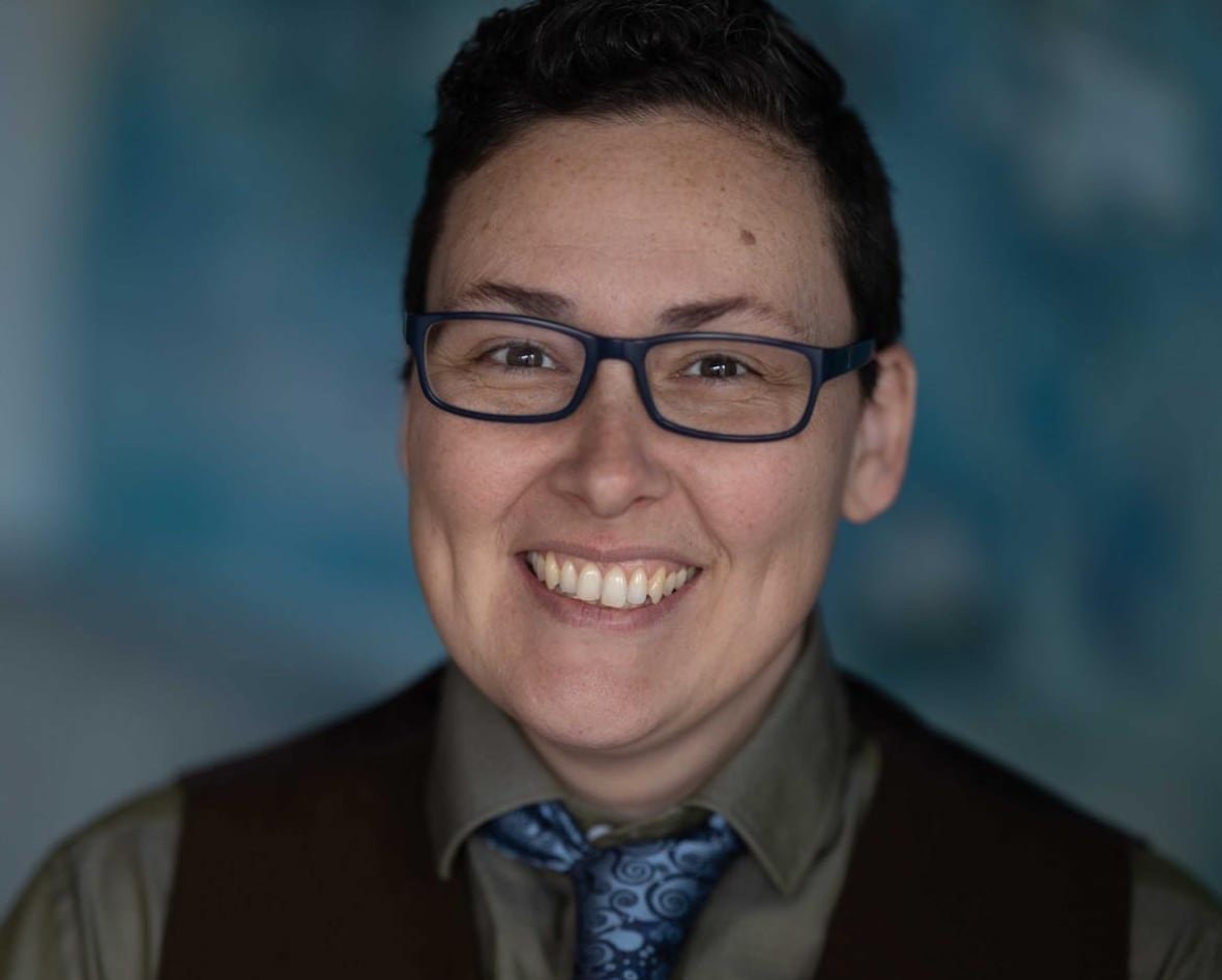 benny-michaud A person with cropped dark brown hair and rectangular glasses, wearing a black vest over a grey button-up shirt and blue patterned tye, smiles brightly for the camera.