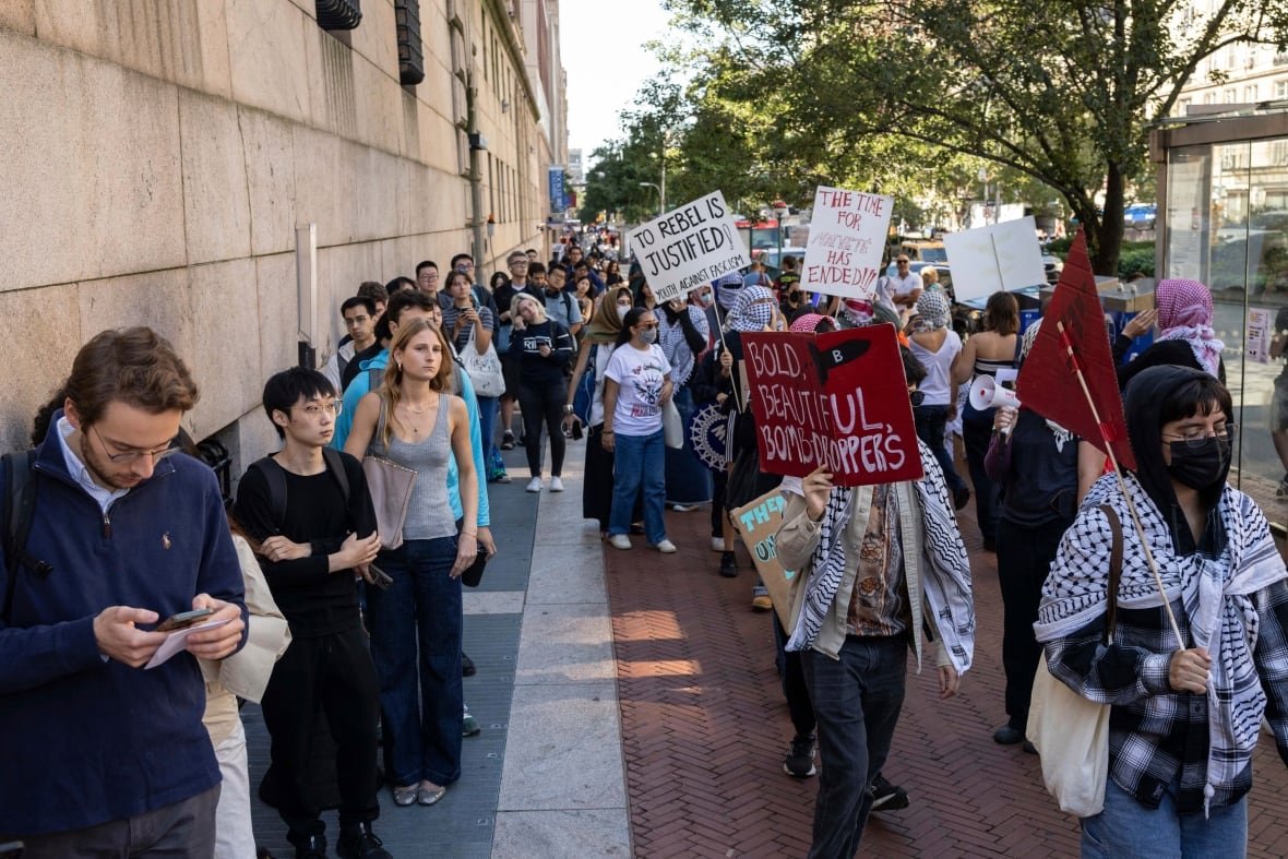 Protesters carrying signs walk past students waiting next to a building.