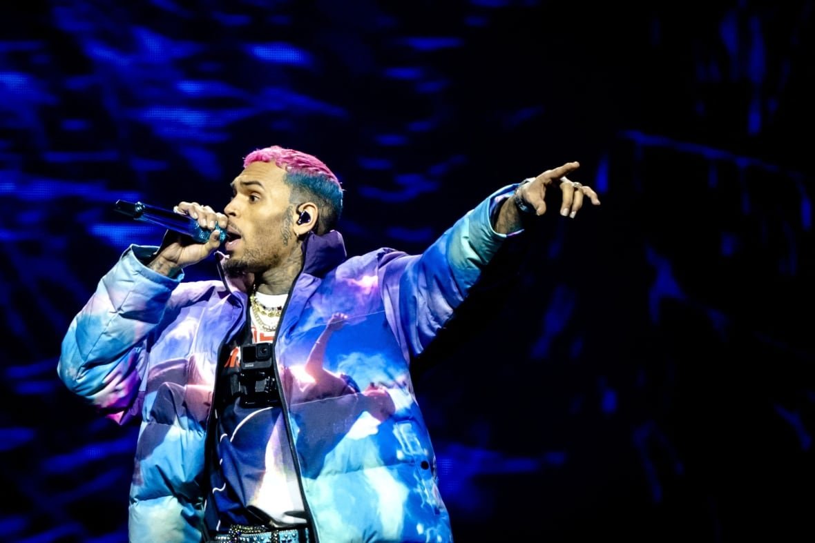 chris-brown-amsterdam-2023 A man hair dyed pink and blue and wearing a puffer jacket, with a horizontal image of a man on it, sings on stage with a microphone in his right hand and pointing to the left with his other hand.