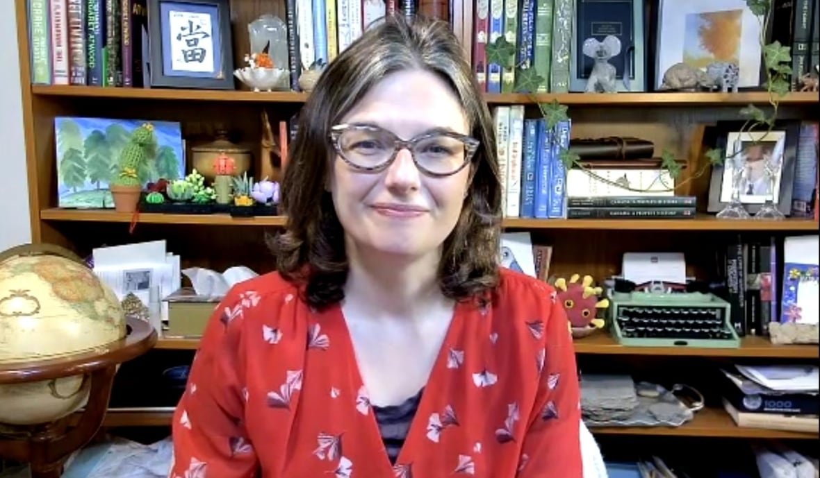 dawn-bowdish-professor-immunology-mcmaster-university-measles A woman with brown shoulder length hair and glasses is wearing a red blouse and sitting in front of a book shelf containing books, plants and a typewriter.