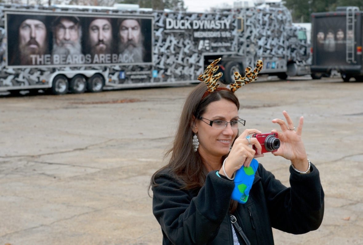 duck-dynasty-hometown A woman wearing leopard-print reindeer ears takes a photo in front of a large tour bus, with a poster bearing the image of four men with long beards.