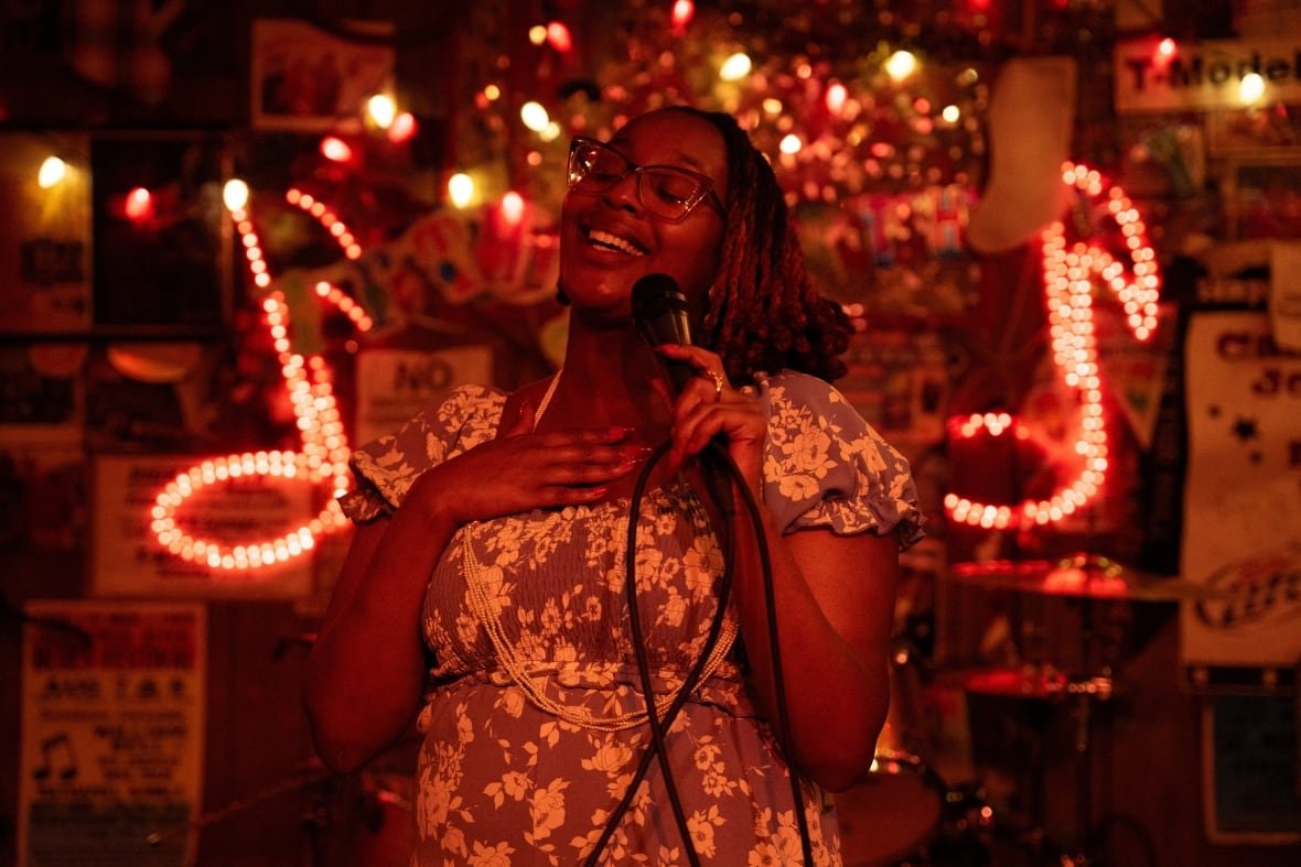 film-sinners-mississippi-1 A woman smiles and sings into a microphone under a red light, her eyes closed and her hand on her heart.