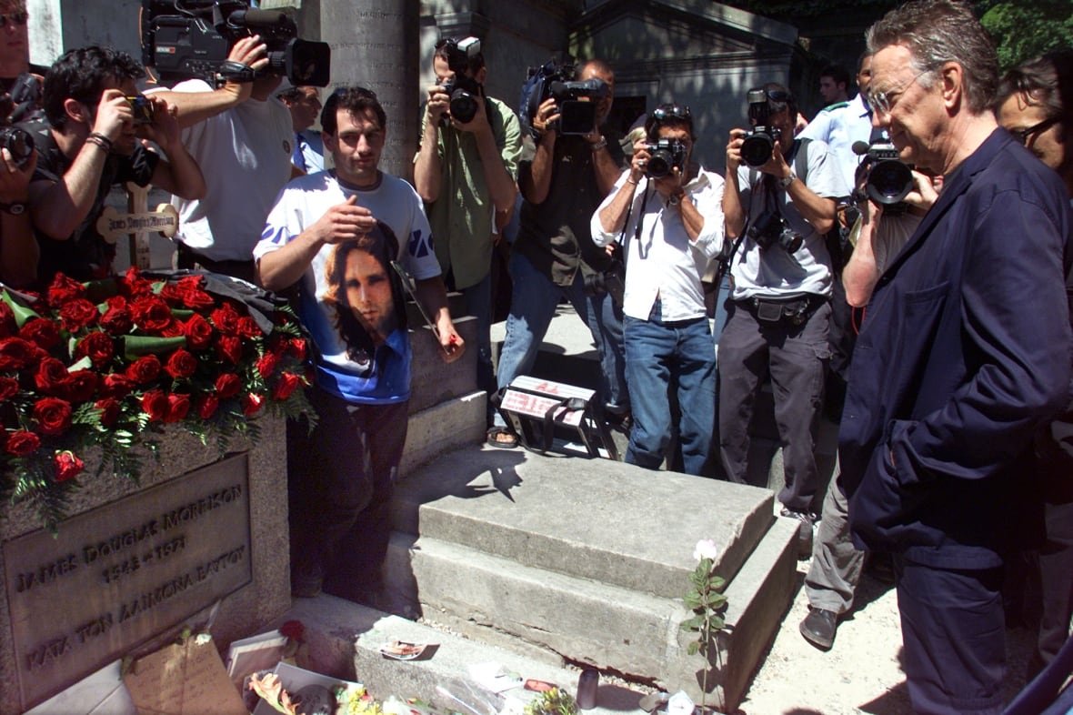 france-ray-manzarek-doors A man in a suit stands in front of a grave stone, covered in red roses, surrounded by onlookers holding cameras.