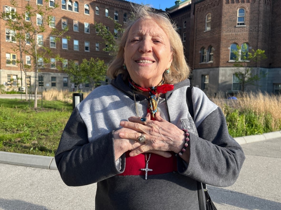 francesca-borrelli An elderly woman wearing a bejeweled cross and rings smiles while holding both her hands to her chest.