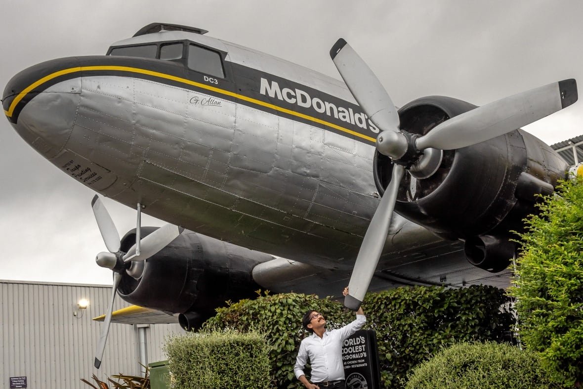 A mustachioed man stands beneath a large silver airplane with the McDonald's logo on it, touching the propeller and looking up.