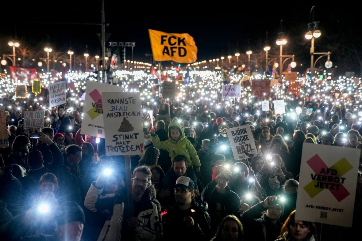 germany-election-protest-far-right A large crowd of people gathered outside at night carry signs and hold up their phones.