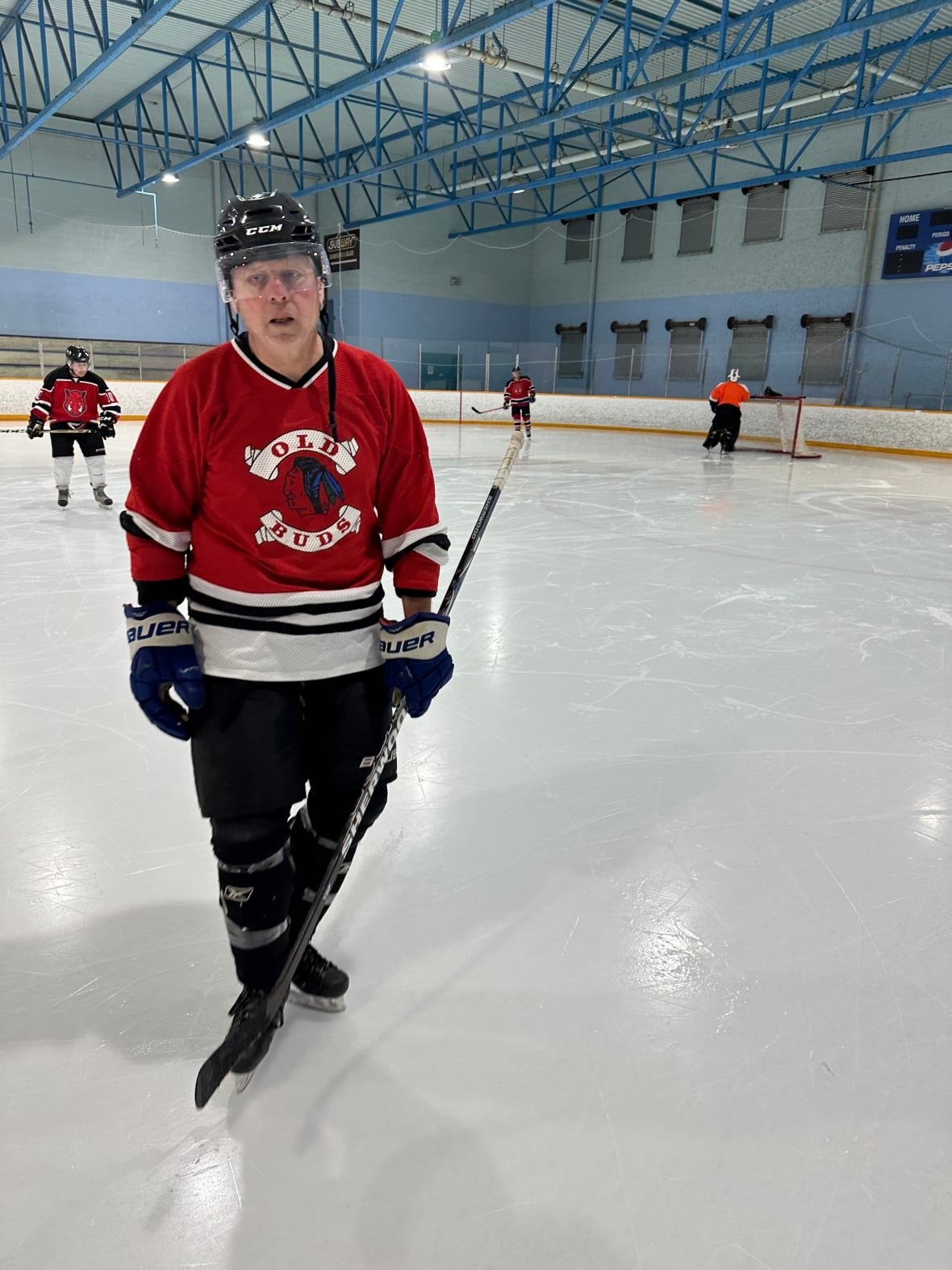 james-smith-playing-hocker A man playing hockey, with other players and a goalie on the rink behind him.