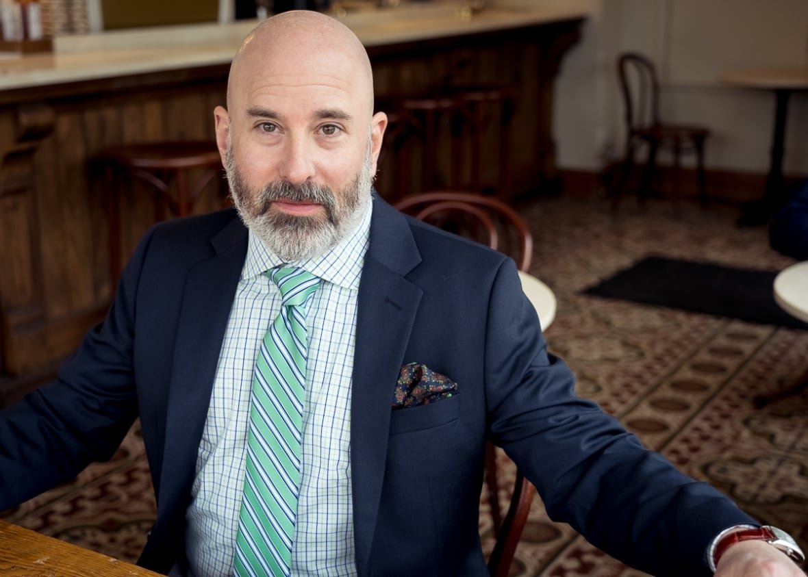 jamie-gruman A man sits at a table in a coffee shop, he is wearing a suit and tie