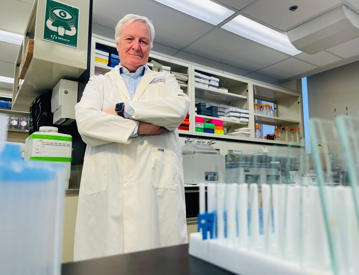 jim-woodgett Man with white hair wearing a lab coat standing in a lab behind test tubes.