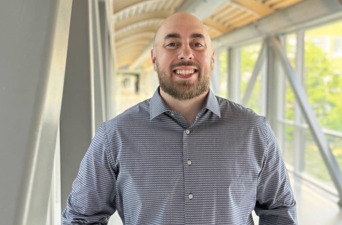 A man in a button-down dress shirt smiles as he stands in a bright indoor walkway with large windows looking over a treed area.