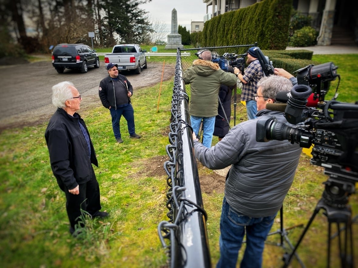 news-camera-point-roberts Journalists with TV cameras mass on one side of a black chain-link fence to interview residents on the other side.