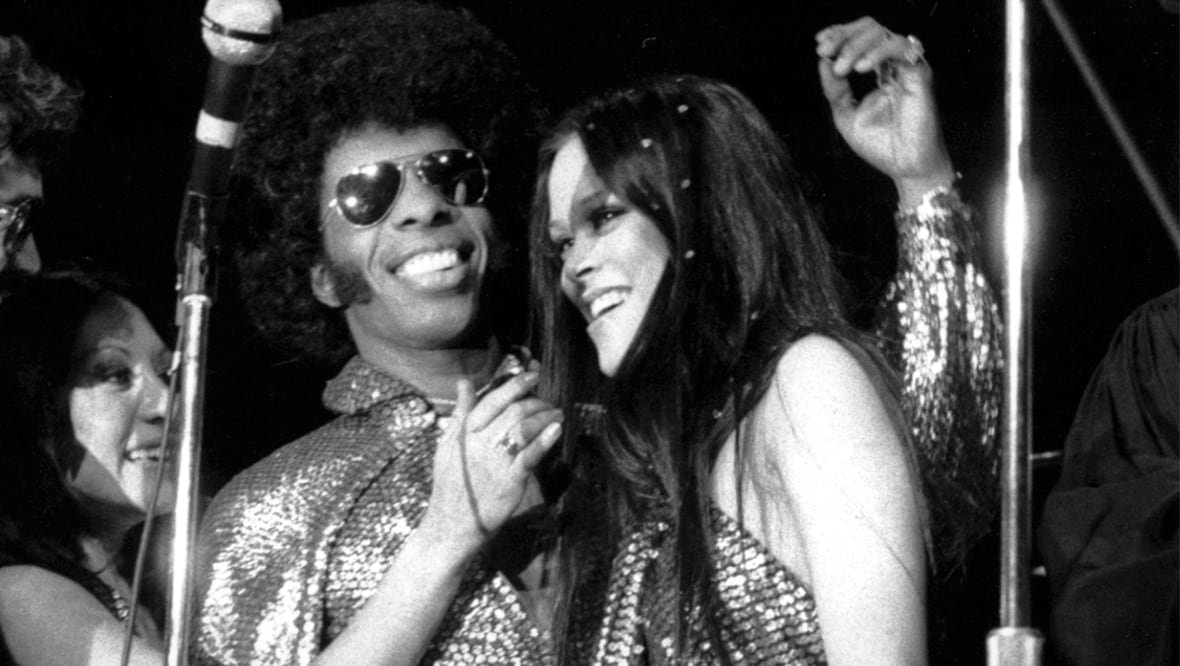 A man smiles with his arm around a smiling woman. He is wearing sunglasses and a number of microphones and mic stands surround them. Another woman at the left side of the image reaches towards the first woman's face.
