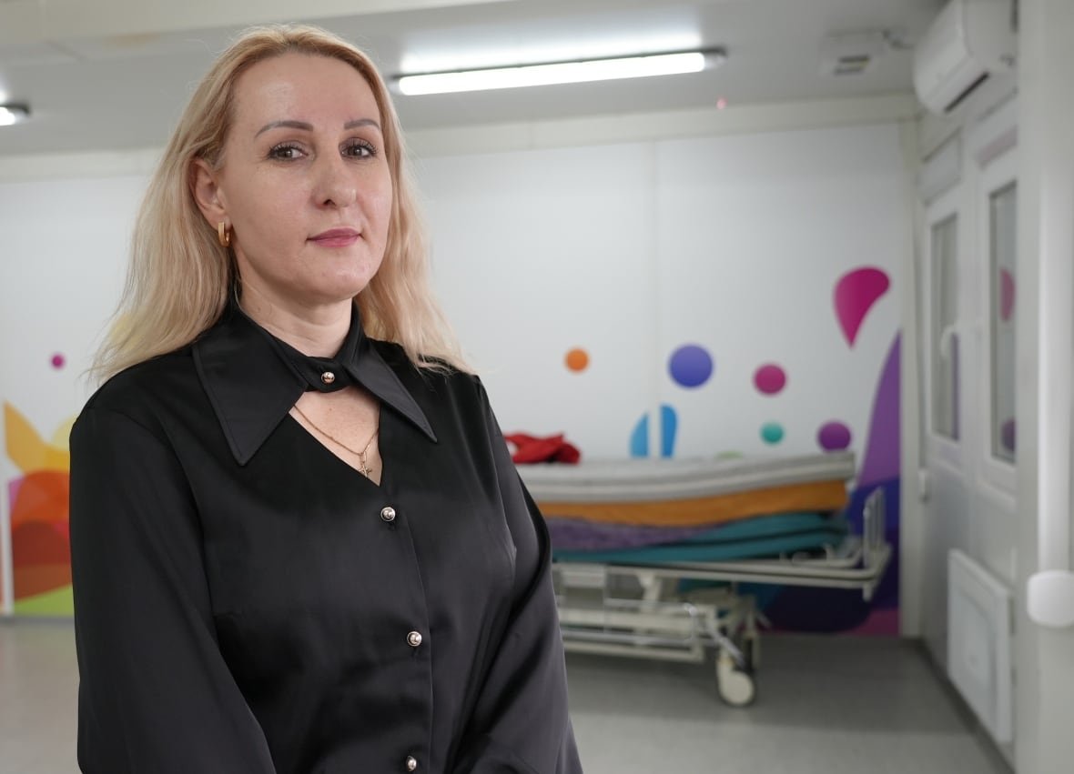olha-tykhomyrova A woman wearing a black blouse poses for a photo while standing in front of a hospital bed pushed against a wall with brightly colours decals on it.
