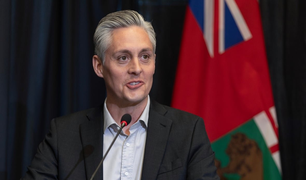 r-j-simpson-premier a man with grey hair wearing a black suit speaks into a microphone in a conference hall with a flag behind him