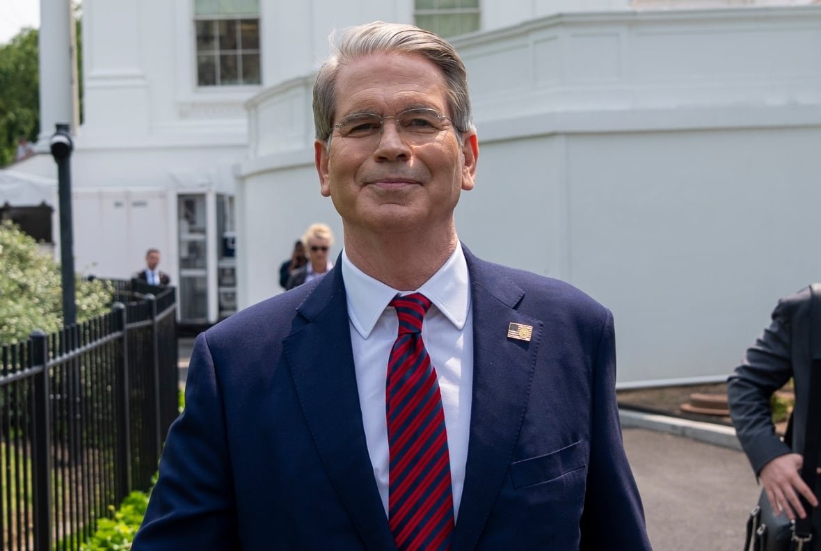 scott-bessent A grey-haired man wearing glasses, a blue suit, and a red and blue striped tie, is shown outside in front of a white building.