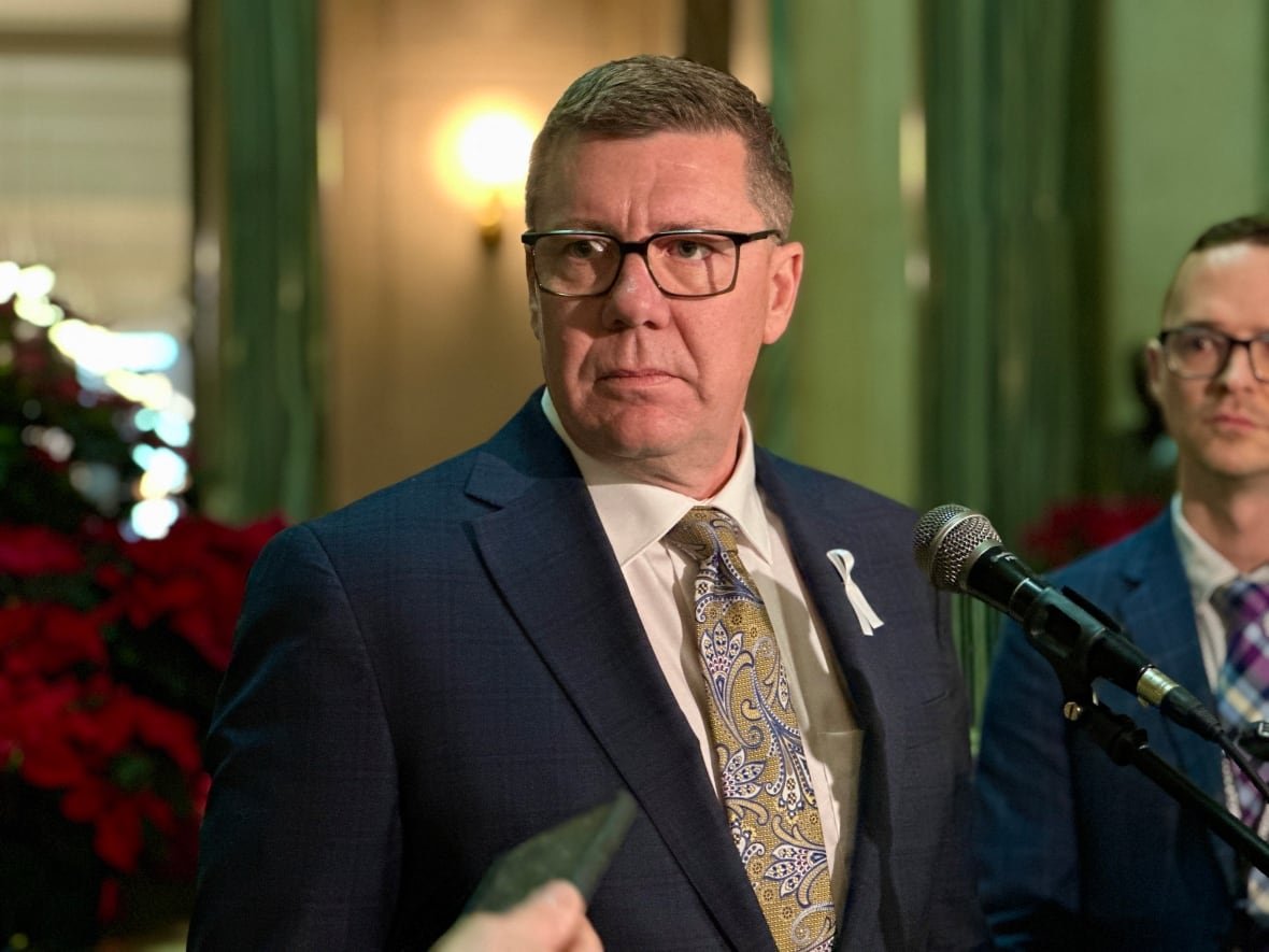 scott-moe A man is wearing a white shirt, blue suit and gold and blue paisley tie. The man is standing in front of a microphone.